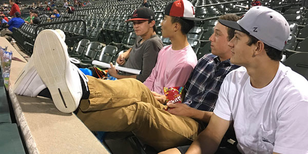 Senior baseball players (left to right) Davis Delich, Eric Yoon, Jacob Daddario, and Mark Simpson sitting along the third baseline taking in a Rough Riders game. 