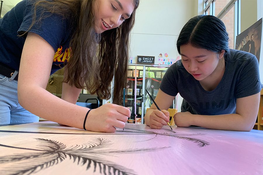 Junior Raina Kinneer and Sophomore Anica Liu paint palm tree leaves onto one of the library tables. The students and their other group members created a pastel beach sunset themed library table. 