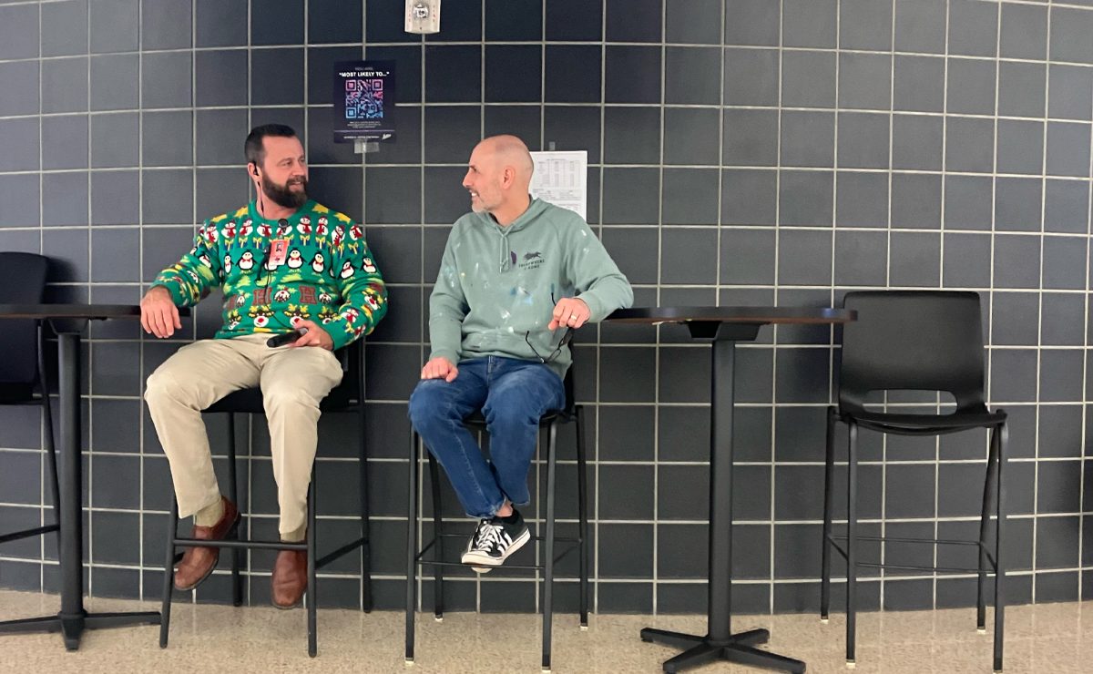 Sitting on the cafeteria stage, assistant principal Jason Harris does his holiday sweater while talking to counselor Ryan Kiefer. 