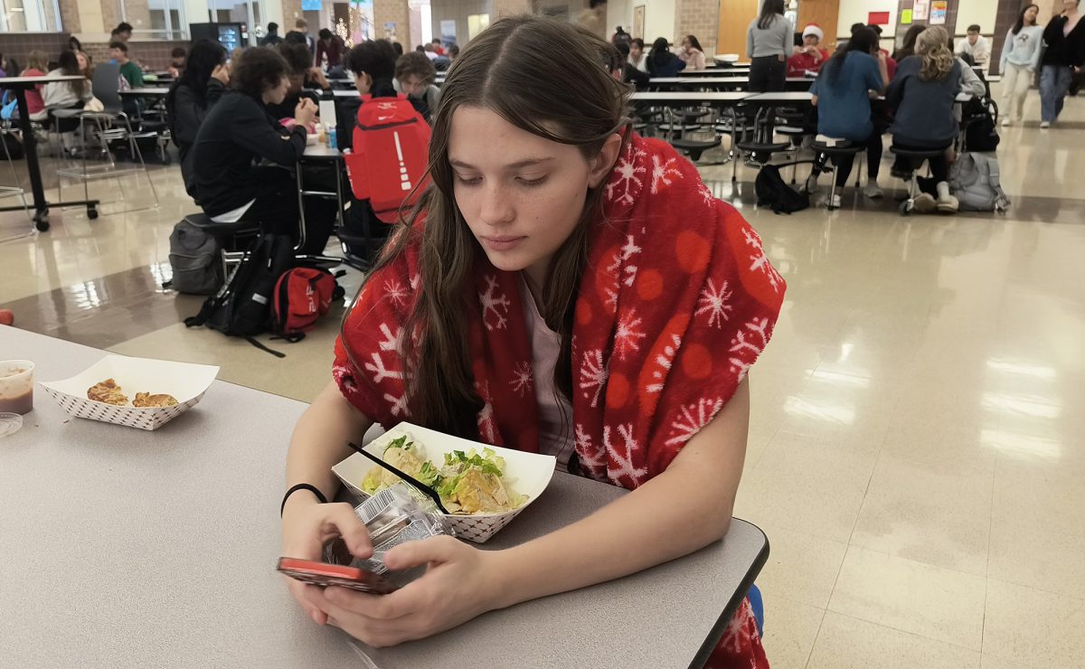 Wrapped in a red blanket with snow flakes, freshman Daisy Demers looks at her phone during lunch on Tuesday. 